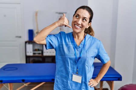Téléchargez les photos : Jeune femme hispanique portant un uniforme de physiothérapeute debout à la clinique souriant faisant geste téléphonique avec la main et les doigts comme parler au téléphone. communiquer les concepts. - en image libre de droit