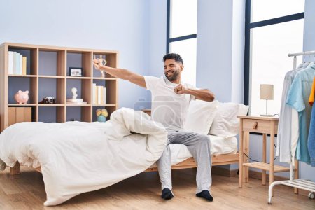 Téléchargez les photos : Young hispanic man waking up stretching arms sitting on bed at bedroom - en image libre de droit