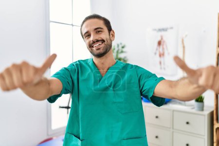 Foto de Young physiotherapist working at pain recovery clinic approving doing positive gesture with hand, thumbs up smiling and happy for success. winner gesture. - Imagen libre de derechos