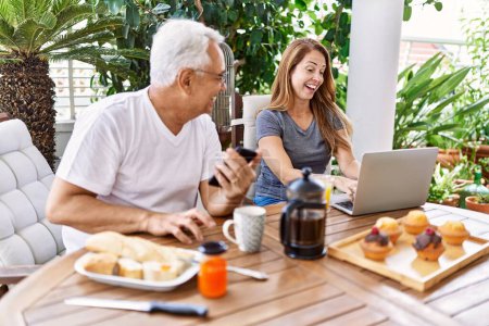 Photo for Middle age hispanic couple having breakfast using smartphone and laptop at the terrace. - Royalty Free Image