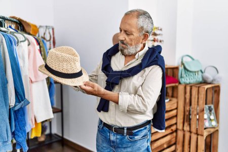 Téléchargez les photos : Homme âgé aux cheveux gris souriant confiant tenant chapeau au magasin de vêtements - en image libre de droit