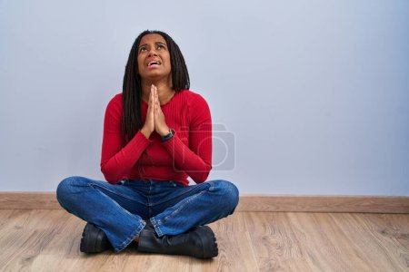 Téléchargez les photos : Jeune afro-américain avec des tresses assis sur le sol à la maison mendier et prier avec les mains ainsi que l'expression de l'espoir sur le visage très émotionnel et inquiet. mendier. - en image libre de droit