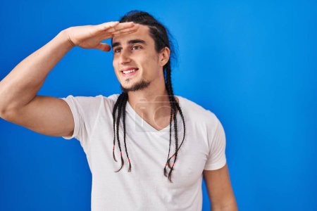 Photo for Hispanic man with long hair standing over blue background very happy and smiling looking far away with hand over head. searching concept. - Royalty Free Image