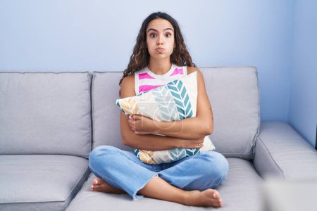 Foto de Joven mujer hispana sentada en el sofá en casa hinchando mejillas con cara graciosa. boca inflada con aire, captación de aire. - Imagen libre de derechos