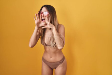 Photo for Young hispanic woman wearing bikini over yellow background shouting angry out loud with hands over mouth - Royalty Free Image