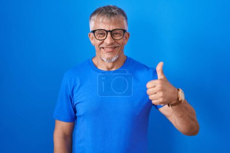 Téléchargez les photos : Homme hispanique avec les cheveux gris debout sur fond bleu faisant des pouces heureux geste avec la main. approuvant l'expression regardant la caméra montrant le succès. - en image libre de droit