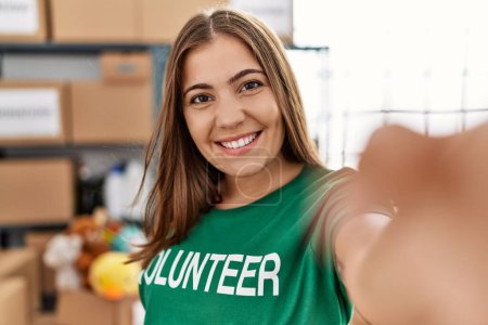 Téléchargez les photos : Jeune femme hispanique portant l'uniforme bénévole faisant selfie par la caméra au centre de charité - en image libre de droit
