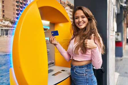 Téléchargez les photos : Jeune femme hispanique prenant de l'argent de guichet automatique avec la carte de crédit souriant heureux et positif, pouce levé faire excellent et signe d'approbation - en image libre de droit