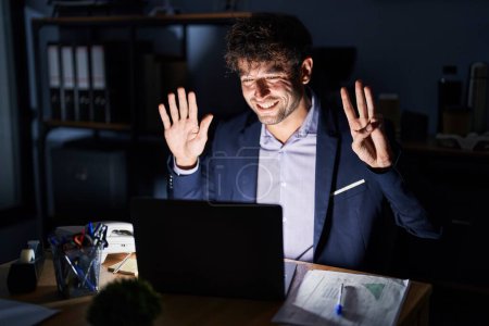 Photo for Hispanic young man working at the office at night showing and pointing up with fingers number eight while smiling confident and happy. - Royalty Free Image