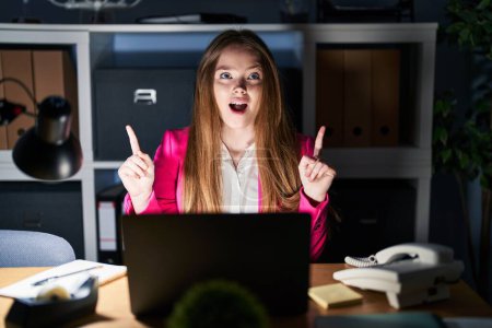 Téléchargez les photos : Jeune femme caucasienne travaillant au bureau la nuit étonnée et surprise regardant vers le haut et pointant avec les doigts et les bras levés. - en image libre de droit
