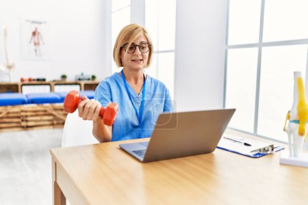 Photo for Middle age blonde woman wearing physio therapy uniform doing tele rehab using dumbbells at clinic - Royalty Free Image