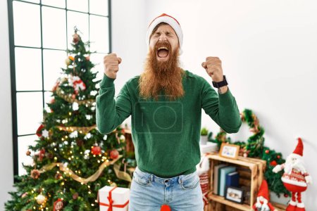 Téléchargez les photos : Homme rousse à longue barbe portant un chapeau de Noël par arbre de Noël excité pour le succès avec les bras levés et les yeux fermés célébrant la victoire en souriant. concept gagnant. - en image libre de droit