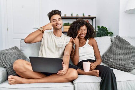 Téléchargez les photos : Jeune couple interracial utilisant un ordinateur portable à la maison assis sur le canapé pointant du doigt vers le visage et le nez, souriant gai. concept de beauté - en image libre de droit