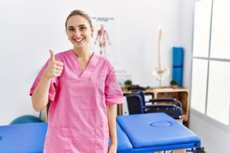 Photo for Young blonde woman working at pain recovery clinic doing happy thumbs up gesture with hand. approving expression looking at the camera showing success. - Royalty Free Image