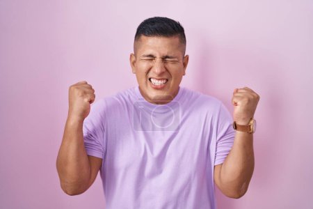 Téléchargez les photos : Jeune homme hispanique debout sur fond rose excité pour le succès avec les bras levés et les yeux fermés célébrant la victoire en souriant. concept gagnant. - en image libre de droit