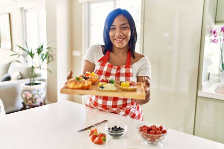 Foto de Mujer morena hispana sosteniendo bandeja con pasteles con frutas en la cocina - Imagen libre de derechos