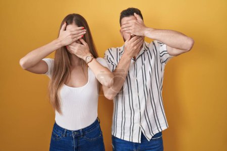 Photo for Young couple standing over yellow background covering eyes and mouth with hands, surprised and shocked. hiding emotion - Royalty Free Image