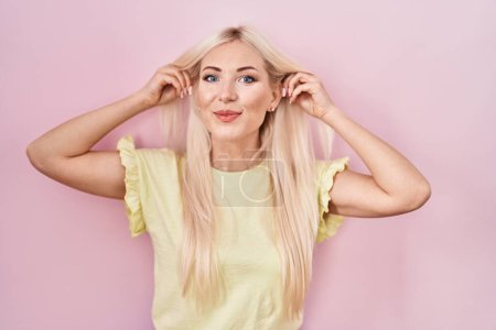 Foto de Caucasian woman standing over pink background smiling pulling ears with fingers, funny gesture. audition problem - Imagen libre de derechos