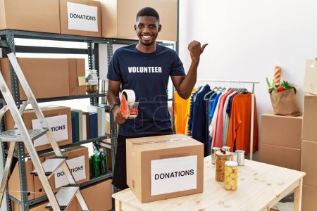 Téléchargez les photos : Young african american volunteer man packing donations box for charity pointing to the back behind with hand and thumbs up, smiling confident - en image libre de droit