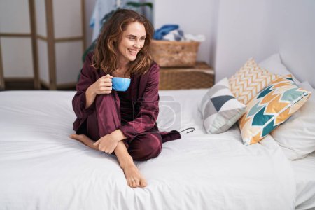 Foto de Mujer joven bebiendo taza de café sentado en la cama en el dormitorio - Imagen libre de derechos