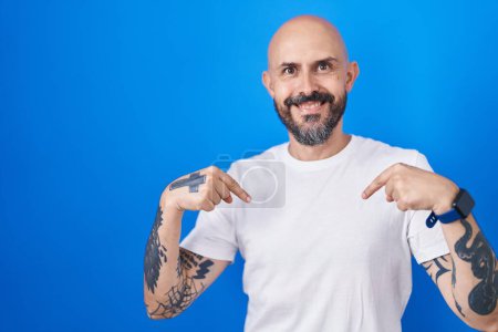 Foto de Hombre hispano con tatuajes de pie sobre fondo azul mirando confiado con sonrisa en la cara, señalándose con los dedos orgullosos y felices. - Imagen libre de derechos