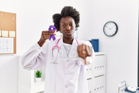 Photo for African doctor woman holding purple ribbon awareness at medical clinic pointing with finger to the camera and to you, confident gesture looking serious - Royalty Free Image