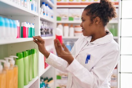 Foto de African american woman pharmacist smiling confident holding toothpaste of shelving at pharmacy - Imagen libre de derechos