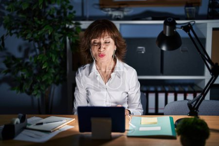 Foto de Mujer de mediana edad que trabaja en la oficina por la noche mirando a la cámara soplando un beso en el aire siendo encantador y sexy. expresión de amor. - Imagen libre de derechos