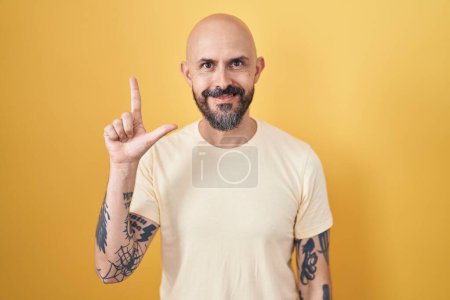 Téléchargez les photos : Homme hispanique avec des tatouages debout sur fond jaune montrant et pointant vers le haut avec les doigts numéro deux tout en souriant confiant et heureux. - en image libre de droit