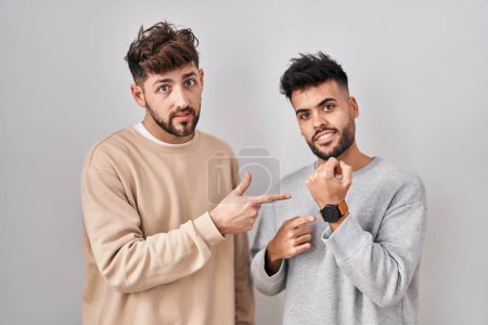 Téléchargez les photos : Young homosexual couple standing over white background in hurry pointing to watch time, impatience, looking at the camera with relaxed expression - en image libre de droit