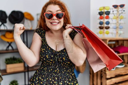 Photo for Young redhead woman holding shopping bags at retail shop screaming proud, celebrating victory and success very excited with raised arm - Royalty Free Image