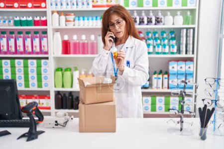 Téléchargez les photos : Middle age woman pharmacist talking on smartphone holding pills bottle at pharmacy - en image libre de droit