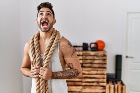 Photo for Young hispanic man training with battle rope at the gym angry and mad screaming frustrated and furious, shouting with anger looking up. - Royalty Free Image