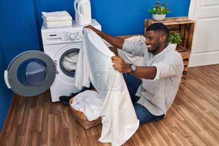 Foto de Joven afroamericano hombre sonriendo lavando ropa en la sala de lavandería - Imagen libre de derechos