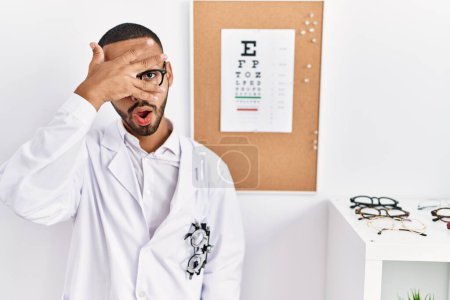Foto de African american optician man standing by eyesight test peeking in shock covering face and eyes with hand, looking through fingers with embarrassed expression. - Imagen libre de derechos