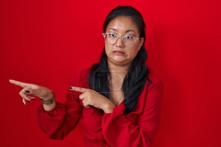 Foto de Asian young woman standing over red background pointing aside worried and nervous with both hands, concerned and surprised expression - Imagen libre de derechos