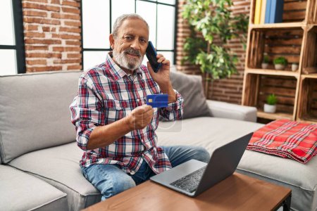 Photo for Senior grey-haired man talking on smartphone holding credit card sitting on sofa at home - Royalty Free Image