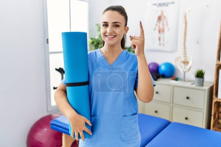 Photo for Young physiotherapist woman holding yoga mat surprised with an idea or question pointing finger with happy face, number one - Royalty Free Image
