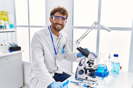 Téléchargez les photos : Jeune homme hispanique portant l'uniforme de scientifique utilisant le microscope au laboratoire - en image libre de droit