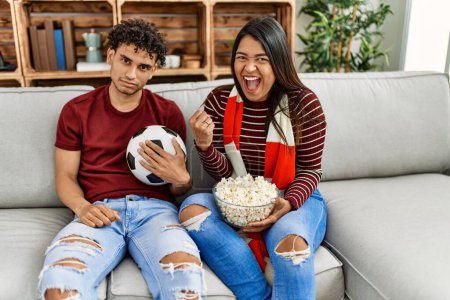 Photo for Young latin couple watching and supporting football match sitting on the sofa at home. - Royalty Free Image