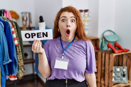 Téléchargez les photos : Jeune rousse femme tenant bannière avec texte ouvert au magasin de détail effrayé et étonné avec bouche ouverte pour surprise, visage d'incrédulité - en image libre de droit