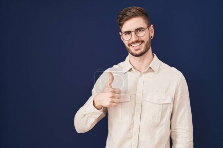 Foto de Hispanic man with beard standing over blue background doing happy thumbs up gesture with hand. approving expression looking at the camera showing success. - Imagen libre de derechos