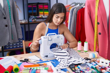 Téléchargez les photos : Jeune belle femme hispanique tailleur en utilisant une machine à coudre au magasin de tailleur - en image libre de droit