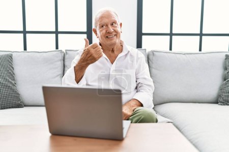 Téléchargez les photos : Homme âgé utilisant un ordinateur portable à la maison assis sur le canapé souriant avec le visage heureux regardant et pointant vers le côté avec le pouce vers le haut. - en image libre de droit