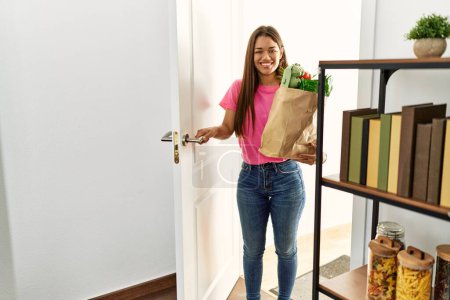 Téléchargez les photos : Jeune femme latine tenant épicerie sac en papier debout à l'entrée de la maison à la maison - en image libre de droit