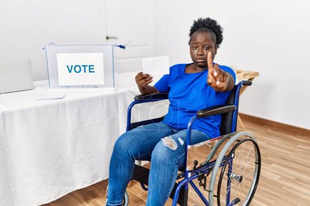 Foto de Mujer africana joven sentada en silla de ruedas votando poniendo sobre en las urnas señalando con el dedo hacia arriba y la expresión enojada, mostrando ningún gesto - Imagen libre de derechos