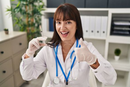 Photo for Young brunette doctor woman holding syringe smiling happy and positive, thumb up doing excellent and approval sign - Royalty Free Image
