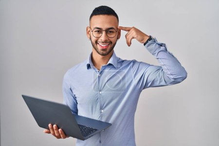 Foto de Joven hombre hispano trabajando usando computadora portátil sonriendo apuntando a la cabeza con un dedo, gran idea o pensamiento, buena memoria - Imagen libre de derechos