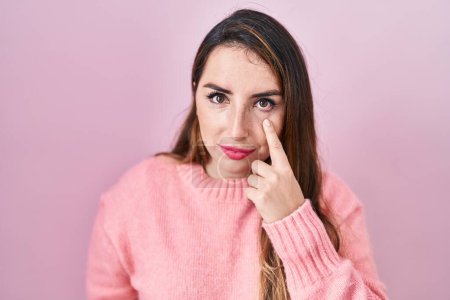Junge hispanische Frau, die über rosa Hintergrund steht und auf das Auge zeigt, das dich beobachtet, Geste, misstrauischer Ausdruck 