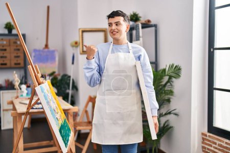 Foto de Non binary person at art studio pointing thumb up to the side smiling happy with open mouth - Imagen libre de derechos